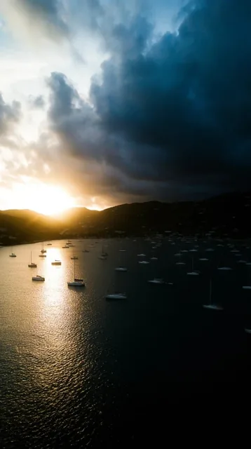A dramatic sunset illuminates a calm bay dotted with dozens of anchored sailboats, framed by da...