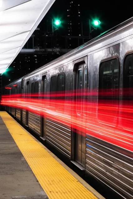 Silver metallic subway train at an outdoor night platform, thick glowing red light trails strea...