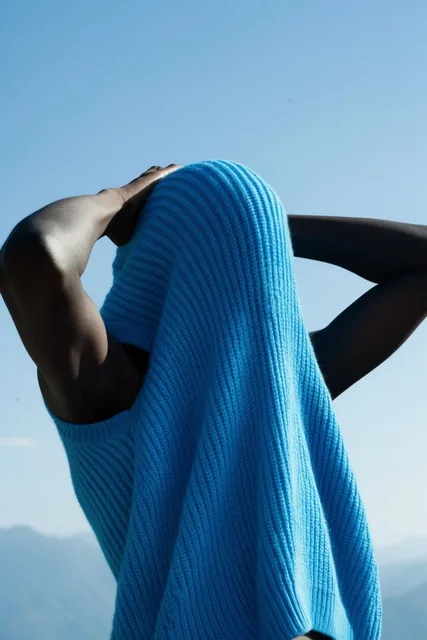 A dark-skinned figure stands with hands clasped behind their head, their face and upper body en...