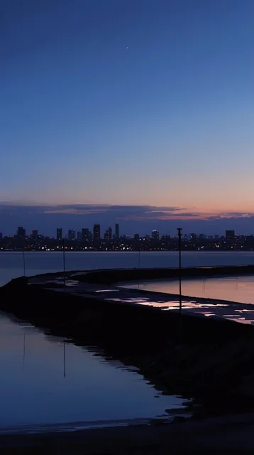 distant city skyline silhouette at dusk, calm reflective water, dark breakwater with glowing we...