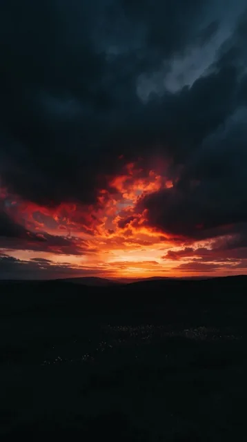 A dramatic sunset bursts through heavy charcoal clouds over a pitch-black rolling landscape. Fi...