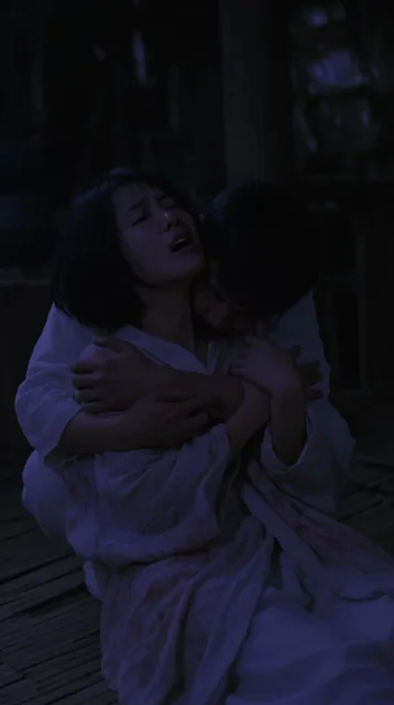 A cinematic medium shot captures a shadowy embrace between a man and a woman in a rustic, dimly...
