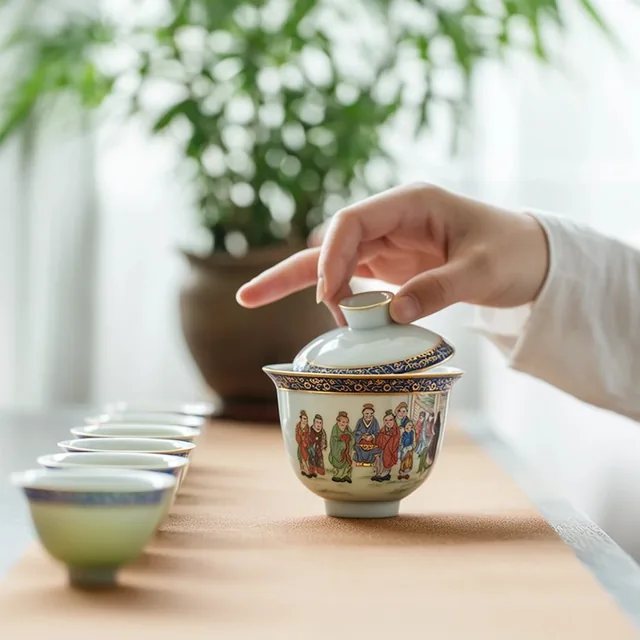hand in white sleeve lifting lid on decorated porcelain gaiwan, painted figures with blue and g...