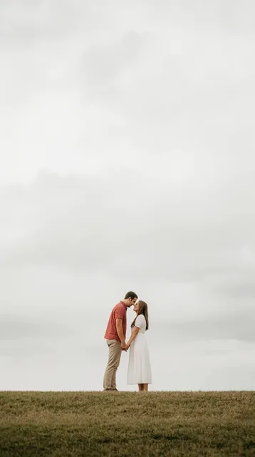 A couple stands face-to-face holding hands on a low grassy horizon beneath a massive, pale gray...