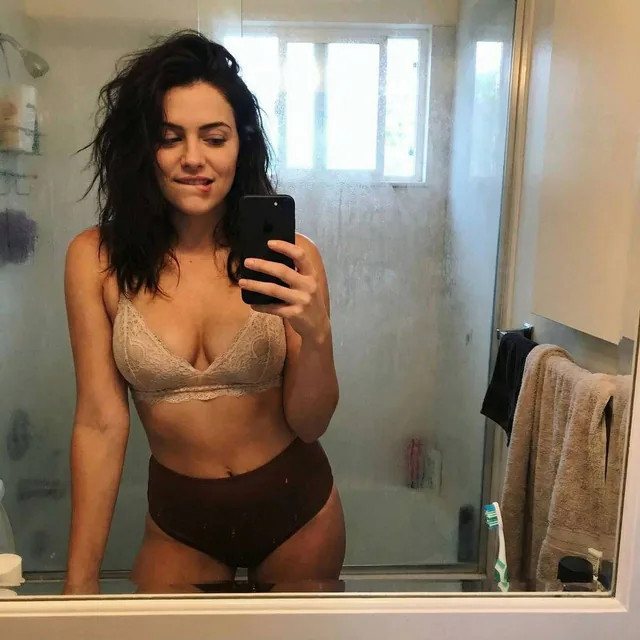 Casual mirror selfie in a bathroom, smartphone photo quality. Slim brunette adult with a...