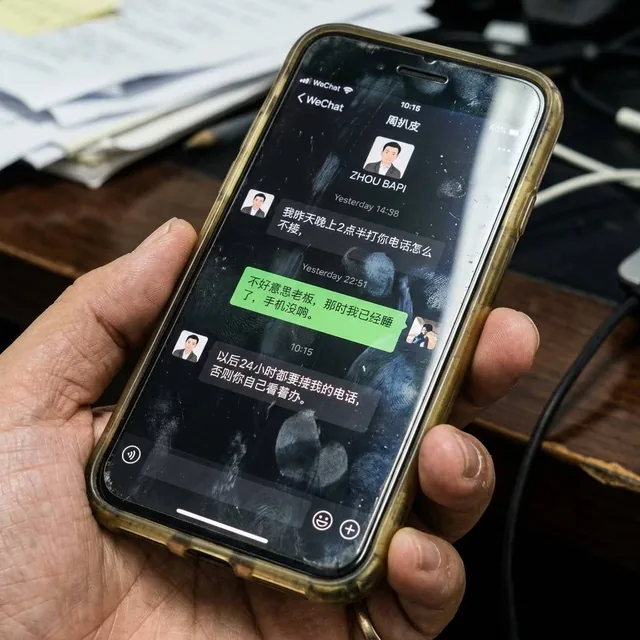 一个手机的屏幕拍摄图，手机屏幕上有手印和反光，手机戴着硅胶套，硅胶套由于用久了有些发黄发黑，屏幕显示内容为微信聊天对话，对方名为周扒皮，对话内容为，周扒皮说，我昨天晚上2点半打你电话怎么不接，微信主人回复不好意思老板，那时我已经睡了，手机没