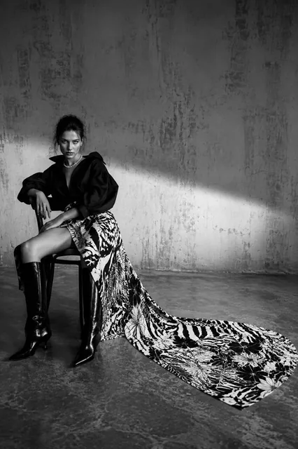 Ultra-realistic black-and-white high-fashion editorial photograph. A model in a dramatic pose sits on a dark wooden chair on the left, wearing an oversized blouse and patterned ski