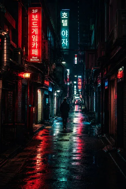 A small, anonymous silhouette of a person in a long coat walks away from the camera down a narrow, wet alley at night, illuminated by red and cyan neon signs reflecting on the pave