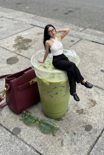 A miniature young woman with long black hair and glasses sits casually on the rim of a large clear plastic cup filled with iced green matcha, wearing a white halter top and black w