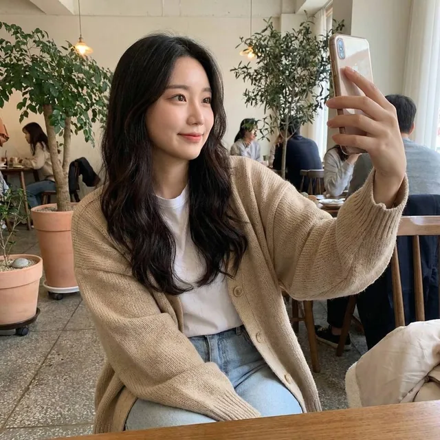 A beautiful young Korean female celebrity in her early 20s, with long black hair and natural Korean makeup, takes a casual smartphone selfie in an everyday setting, exuding a spont