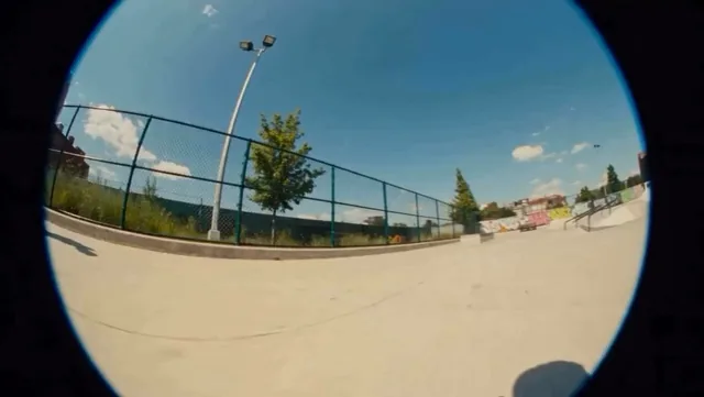 Fisheye lens skatepark shot on a sunny day with chain-link fences and graffiti-covered ramps in the background. Camera s