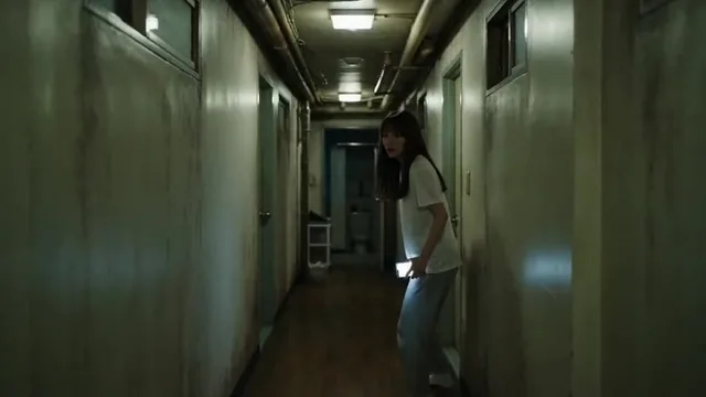 A young woman in pajamas holding a dim phone light walks down a dark, narrow apartment hallway at night, her face tense with fear as she approaches a slightly ajar bathroom door.