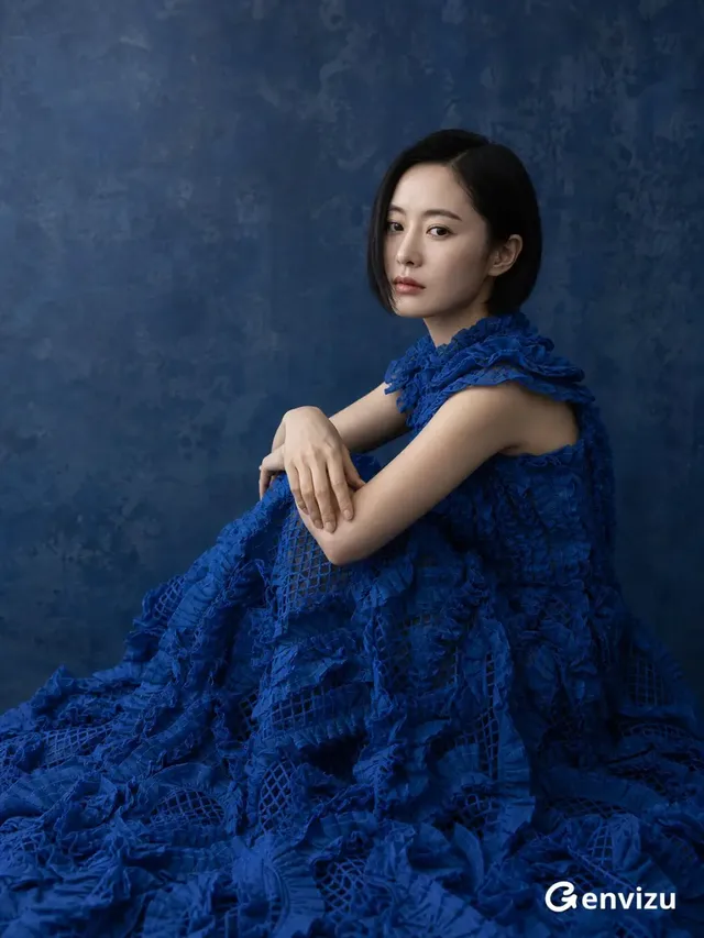 Fine-art fashion portrait of an East Asian woman with a chin-length bob, wearing a voluminous cobalt-blue ruffled couture gown, seated in a curled pose against a mottled indigo studio wall.