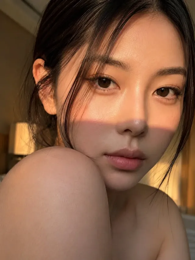 Ultra-realistic close-up beauty portrait of a young East Asian woman looking over her shoulder into the camera during golden hour, with warm light illuminating her face and a softly blurred hotel bedroom background.