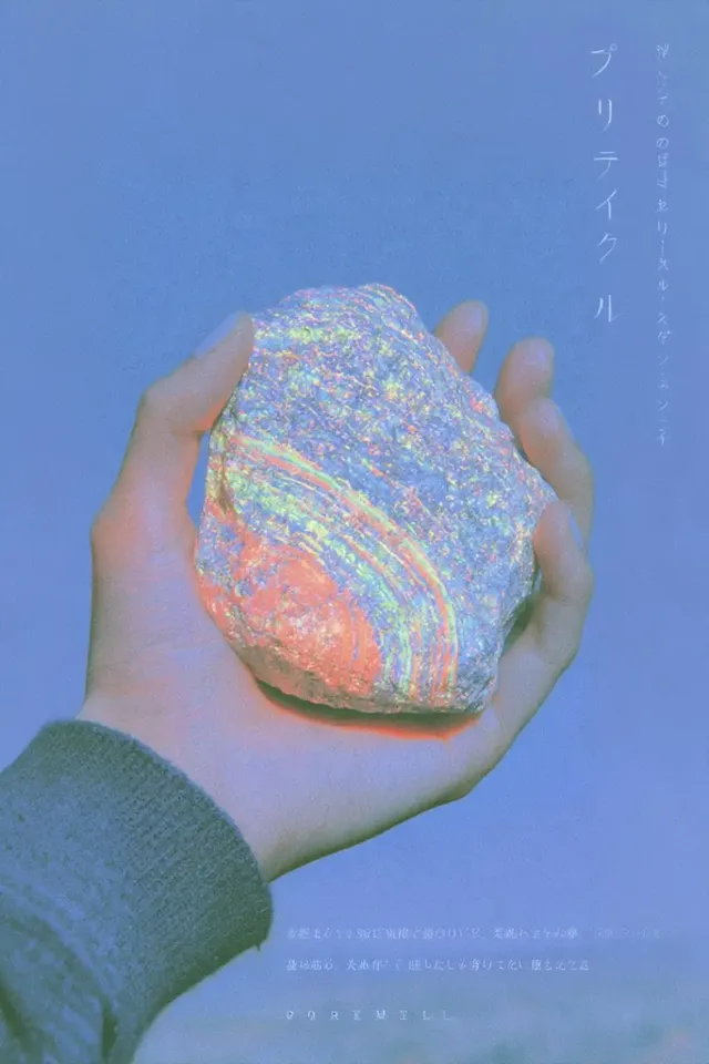 A person's hand holding a rough, grey rock that glows with vibrant neon colors like pink, orange, green, and blue under UV light, set against a plain blue background.