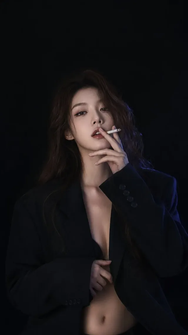 A young woman with long, wavy brown hair and fair skin, wearing a black blazer, sits with her right hand near her lips holding a cigarette, her eyes looking directly at the camera with a subtle allure, highlighted by soft front lighting and cool blue rim lighting on a black background.
