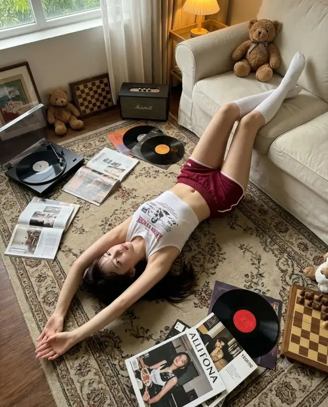 Overhead view of a woman with Korean glass skin lying on a rug in a vintage living room, her body in an S-curve, with an iPhone 17 Pro perspective.