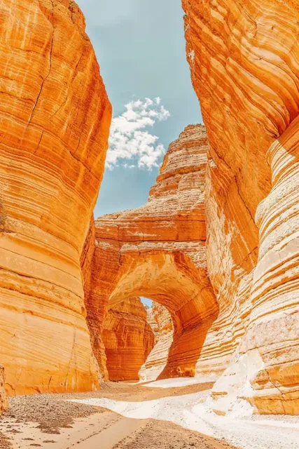 A vibrant desert canyon with layered orange rock formations and a natural arch, under a bright blue sky with white clouds.
