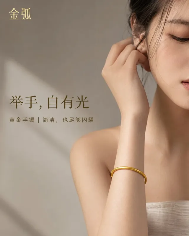 Close-up of a Chinese woman's shoulder, neck, and arm, with her wrist gently placed near her face, showcasing a sleek, minimalist gold bangle. The background is a soft, warm grey, with subtle window lighting highlighting the bracelet.