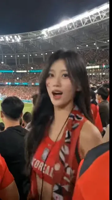 Close-up smartphone video of an excited young East Asian woman with a South Korean flag sticker on her cheek, wearing a red football top and scarf at a night match.