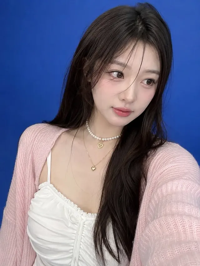 Ultra-realistic smartphone selfie of a beautiful young East Asian woman with dark hair, wearing a white camisole dress, pearl choker with heart pendant, and a pink cardigan, against a seamless blue studio backdrop. She has a gentle expression and is looking slightly away from the camera.