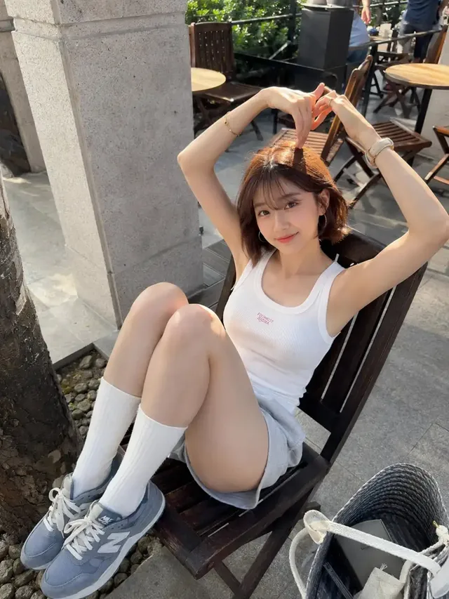 Full body vertical smartphone photo of a young woman sitting in an outdoor cafe chair, forming a slightly asymmetrical heart with her fingers above her head, looking up at the camera. She wears a white tank top, grey shorts, and knee-high socks.