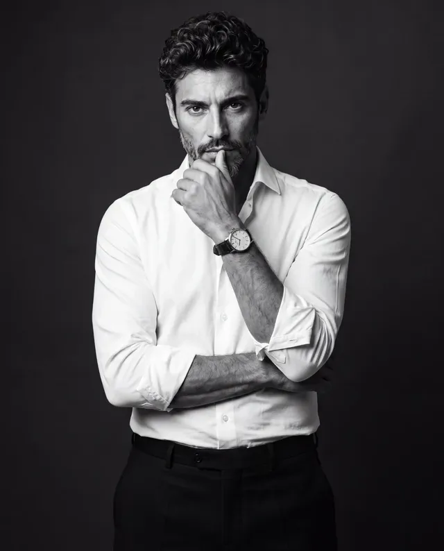 Upper thigh to head studio portrait of a man in a white dress shirt and black trousers, one arm folded, the other hand touching his chin, looking intently at the camera with a sharp expression.