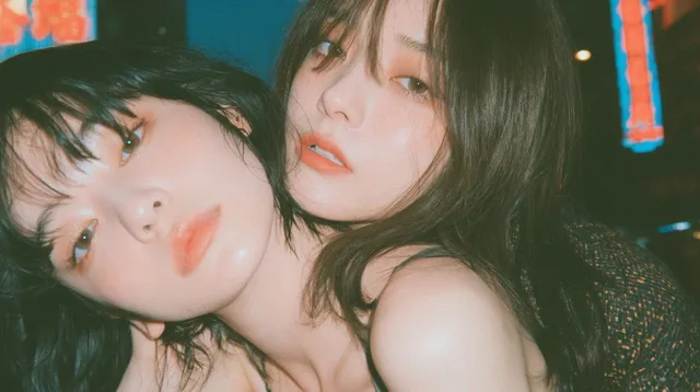 Two young women in an asymmetrical, off-center fashion portrait. One woman is cropped at the edge of the frame, with the other slightly behind her. They are in a crouching pose against a blurred background of bright neon signs and dense street life in Hong Kong's Mong Kok district. Dappled sunlight falls on their faces and shoulders.
