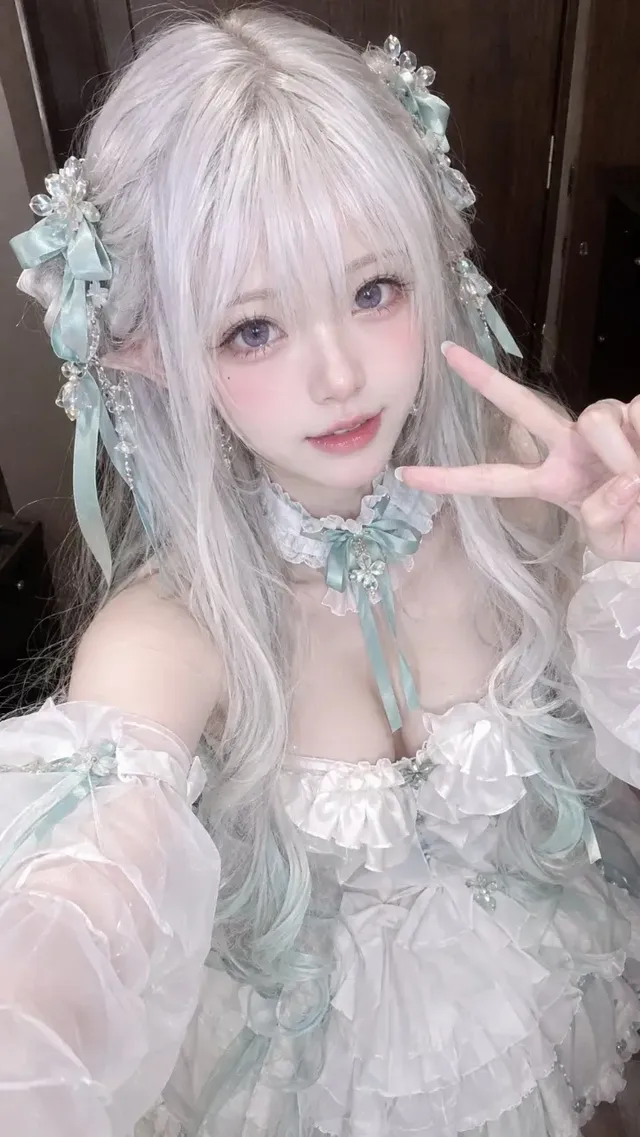 A portrait of a young East Asian female cosplayer with silver-white hair and violet eyes wearing a white princess gown, taking a selfie.