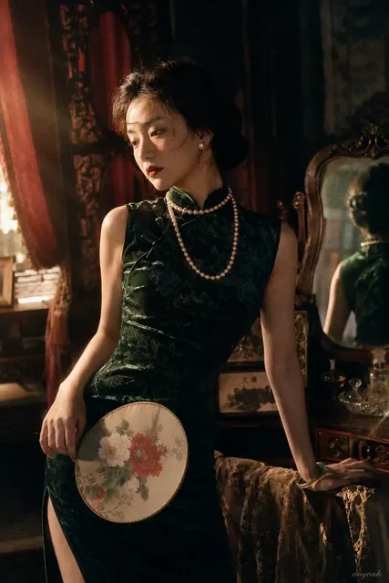 Woman in a dark green qipao and pearl necklace, holding a fan, standing by a window in a dimly lit vintage room.
