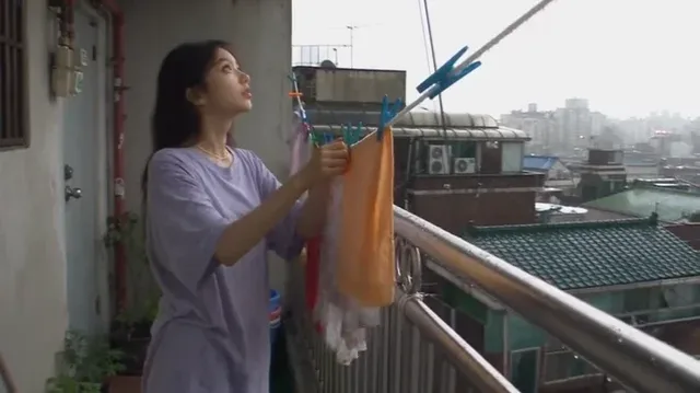 Young Korean woman laughing on a wet apartment balcony in Seoul during a sudden summer rainstorm, filmed with a shaky, low-fidelity early 2000s camcorder.