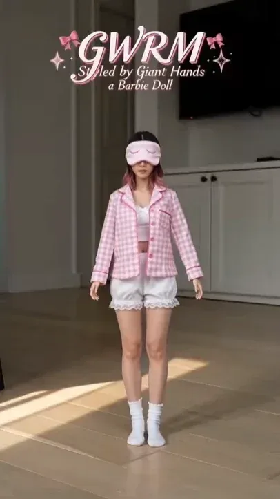 A miniature person styled like a Barbie doll stands on a wooden floor, with enormous hands from off-screen manipulating her clothing and accessories.