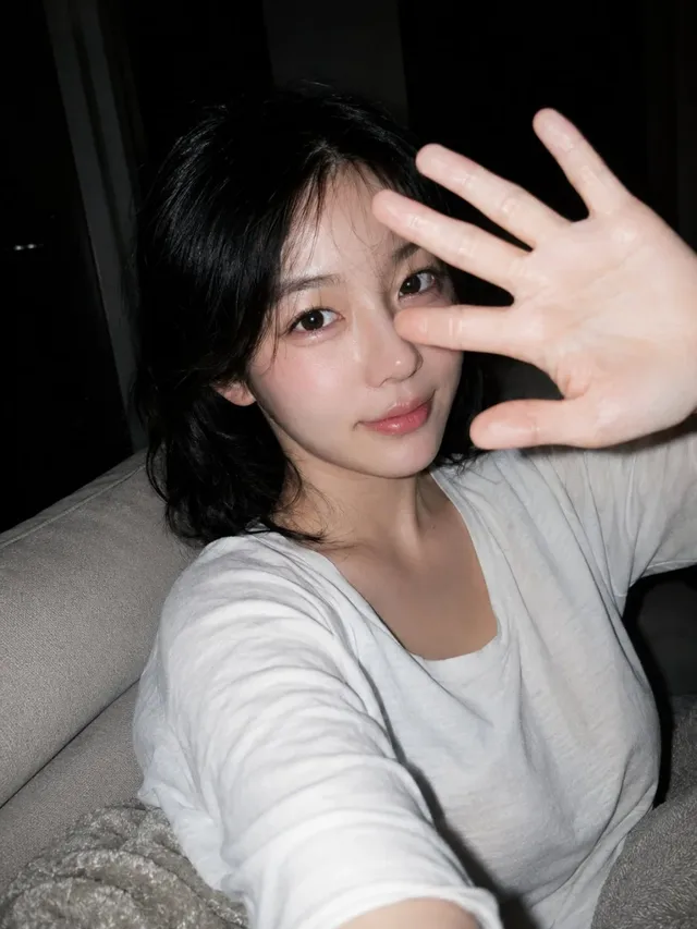 Young East Asian woman with natural makeup and slightly messy black hair, wearing an oversized white T-shirt, looking at the camera with a gentle smile from a home sofa. One hand is near the lens, slightly blurred due to proximity.