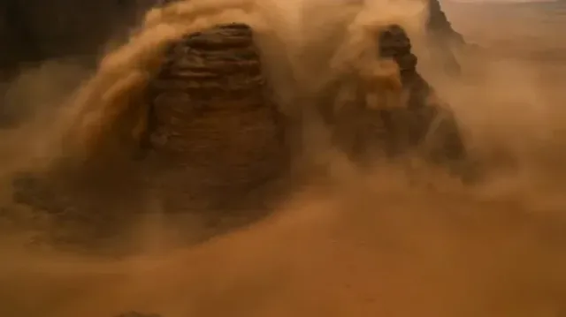 Aerial view of an off-road buggy speeding through a violent sandstorm in a desert, with collapsing cliffs and dunes, before emerging onto a salt flat at sunset.