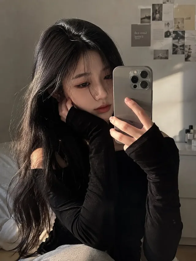 Ultra-realistic smartphone mirror selfie of a beautiful young East Asian woman with black hair, wearing a black off-shoulder sweater, taking a photo of herself in a modern bedroom.