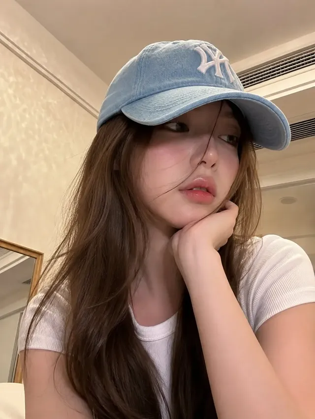 Ultra-realistic smartphone portrait of a young East Asian woman with ash-brown hair, wearing a blue baseball cap and white t-shirt, looking directly at the camera with a relaxed expression in a modern bedroom.