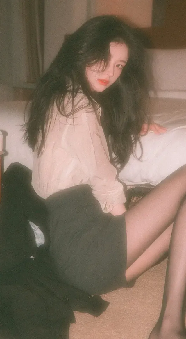 A young Korean female idol sits on a hotel carpet, leaning against the bed, wearing a beige shirt, grey skirt, and black stockings, with a casual, relaxed expression.