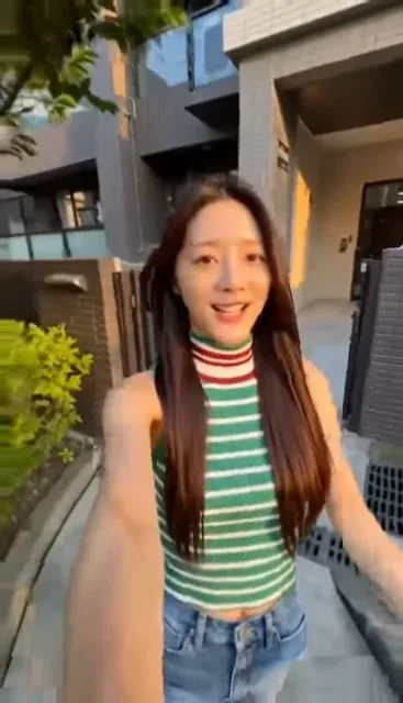 A young Korean K-pop idol with long, dark, straight hair, wearing a green and white striped top and blue jeans, smiles and vlogs with her smartphone during a relaxed day in a Taiwa