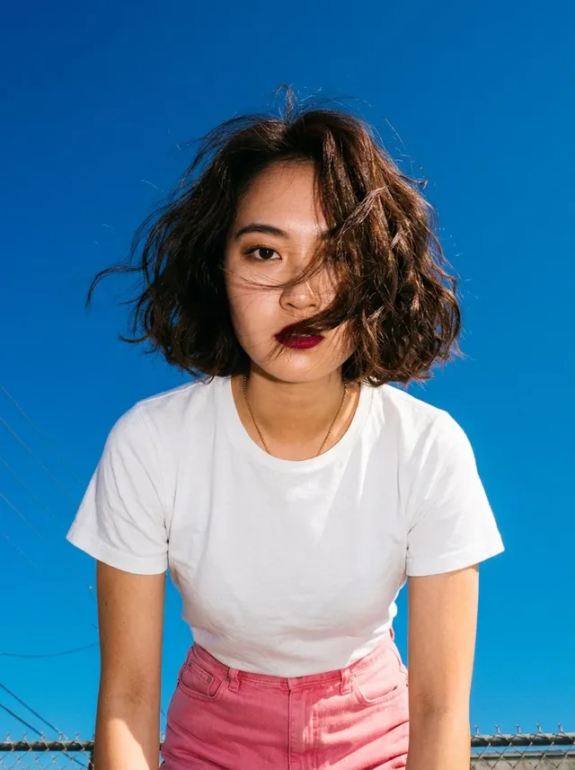 Extreme low-angle shot of a young East Asian woman with a bob haircut and red lipstick, wearing a white t-shirt and pink denim mini skirt, looking directly at the camera with wind-blown hair against a dramatic, saturated electric blue sky.