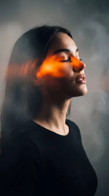 Side profile of a woman with closed eyes and parted lips, bathed in chiaroscuro lighting with vibrant orange highlights and a smoky background.