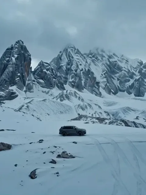 Woman with dark hair wearing a Yankees cap and grey athleisure dancing on the roof of an SUV in a snowy mountain landscape.