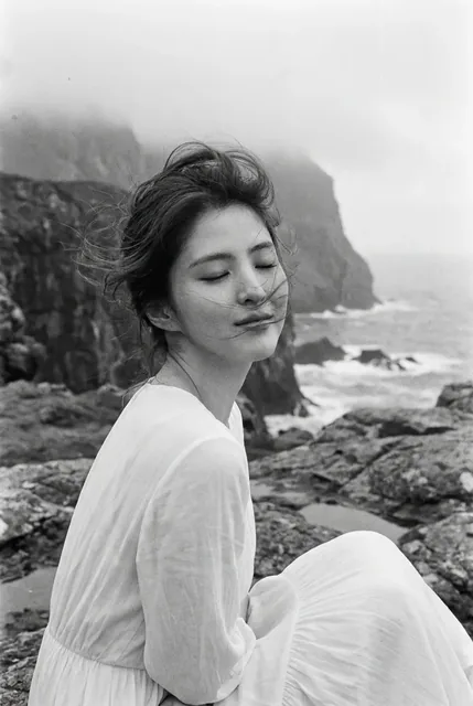 Black and white film photo of a woman with windblown hair and closed eyes, sitting on seaside rocks with a misty cliff in the background.