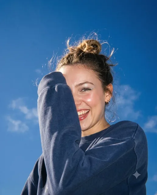 A young woman with a messy bun laughs, shielding her face with her forearm, in a low-angle, ultra-realistic early 2000s digicam photo.