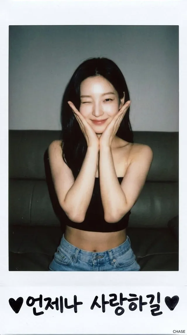 A young woman with dark wavy hair, wearing a black bandeau top and low-rise light blue jeans, poses in a vintage Polaroid frame, winking with a playful smile and a soft focus effect.