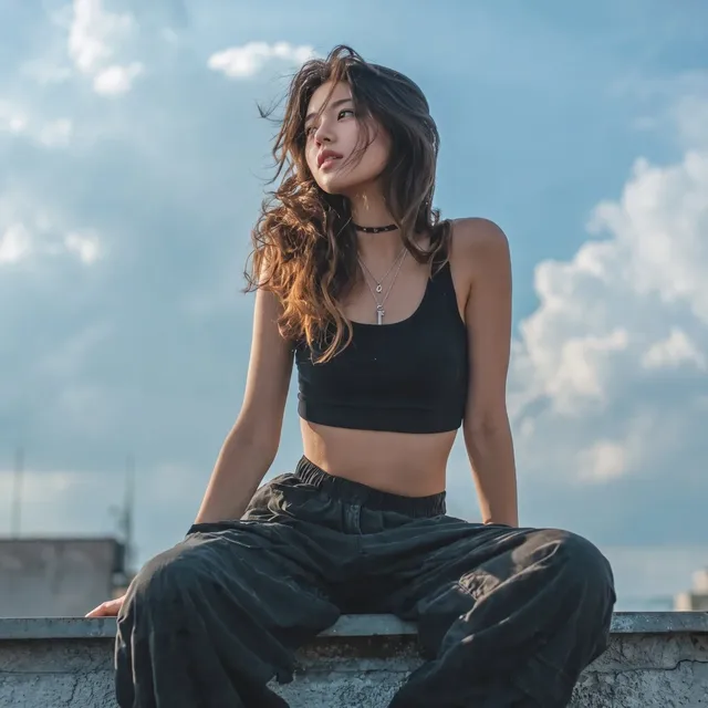 A full-body editorial portrait of a young East Asian woman in a black crop top and cargo pants sitting on a rooftop ledge during golden hour.