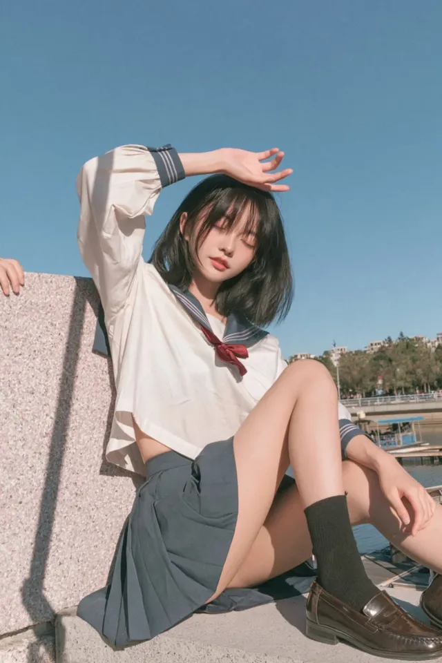 A young Japanese schoolgirl with a black bob haircut and bangs, wearing a white sailor uniform with a red bow, sits on a textured stone wall. She is shielding her eyes from the sun with her hand, and her legs are crossed. The background is a soft blue sky and blurred landscape.