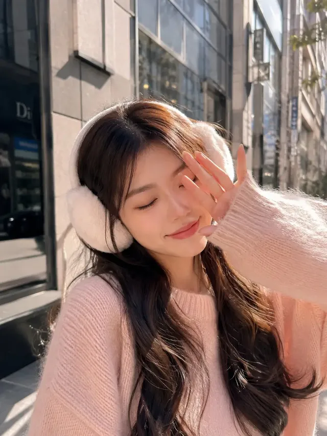 A young woman with long dark hair and closed eyes smiles gently, wearing a pink sweater and white earmuffs, bathed in warm sunlight on a city street with blurred modern buildings.