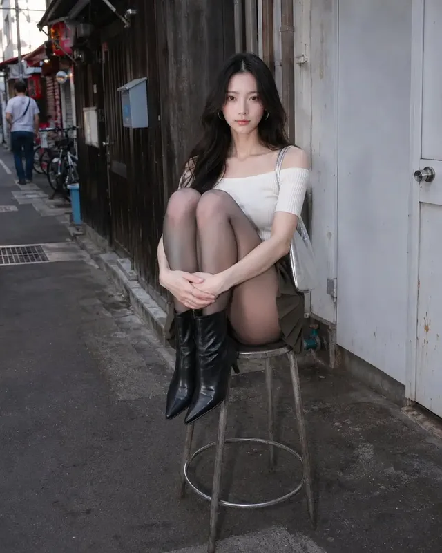 A person wearing a fitted ivory off-shoulder top, dark olive low-rise pleated miniskirt with utility straps, sheer charcoal-gray tights, and black leather ankle boots, with a silver shoulder bag and hoop earrings.