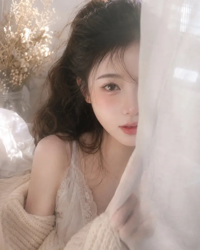 Close-up of a young Korean woman's face, partially obscured by a sheer white veil, with soft window light creating a gentle glow. One eye and part of her lips are visible.
