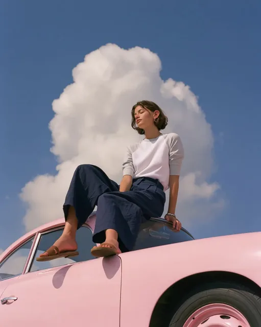 Minimalist young woman with dark bob haircut sitting on a pastel pink vintage Porsche 356, low-angle shot against a vast blue sky with cumulus clouds.